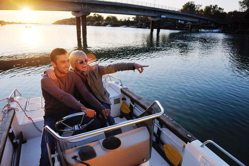 Couple driving a boat at sunset under a bridge, smiling about boat insurance in Frankenmuth, MI